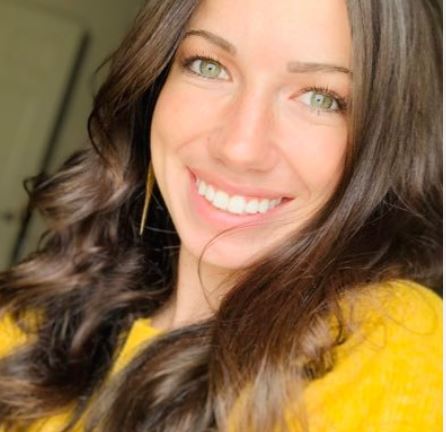 Close-up of a smiling woman with long dark hair wearing a yellow top.