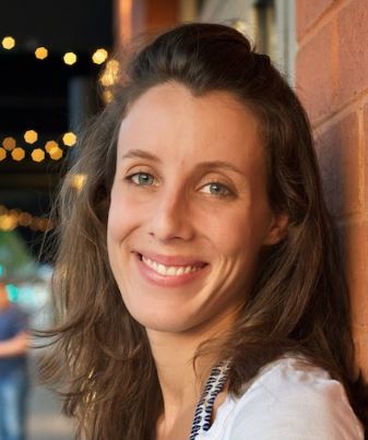 Smiling woman with long brown hair leaning against a brick wall with blurred lights in the background.