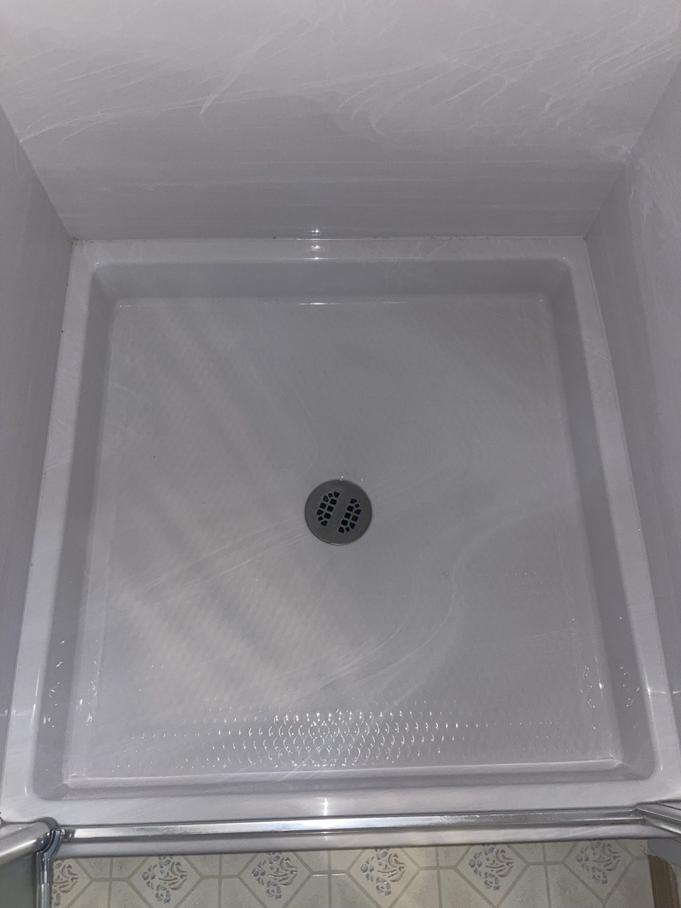 Empty white square shower stall with a central round drain and patterned floor tiles visible outside.