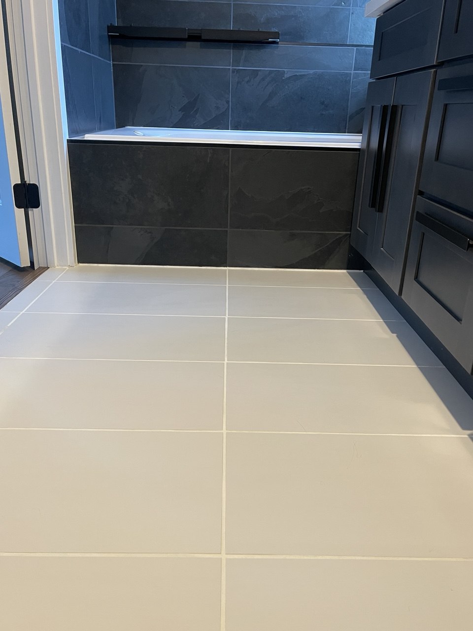 Bathroom floor with large beige tiles, dark tiled bathtub surround, and dark wood cabinetry.