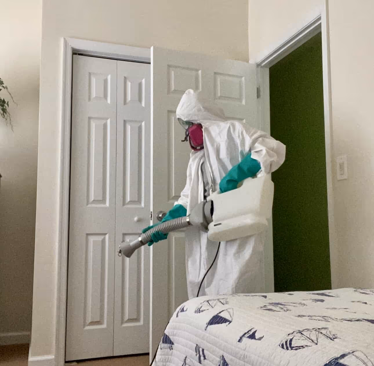 Person in white protective suit, pink respirator mask, and green gloves disinfecting a bedroom with a fogging machine.
