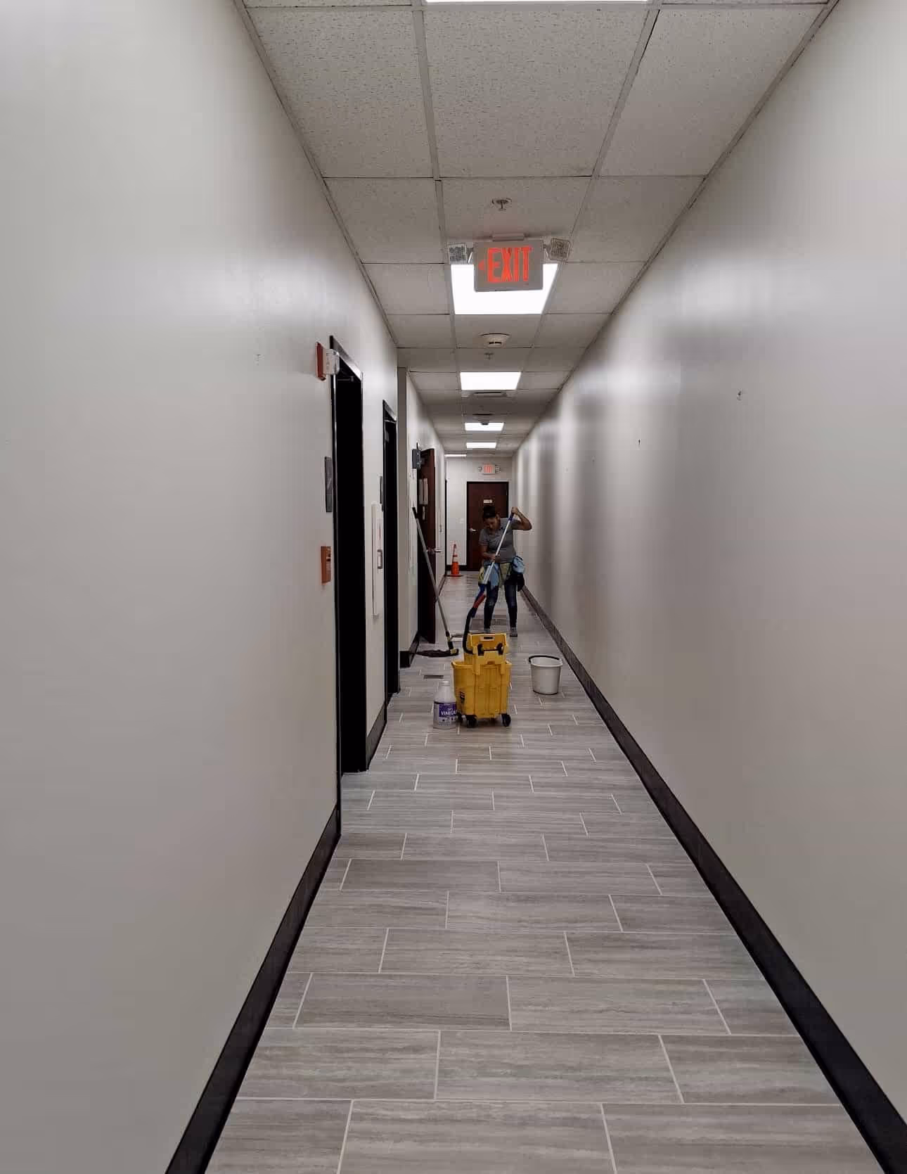 A person mopping a long hallway with white walls and tiled floor, with cleaning supplies including a mop bucket and a white bucket.