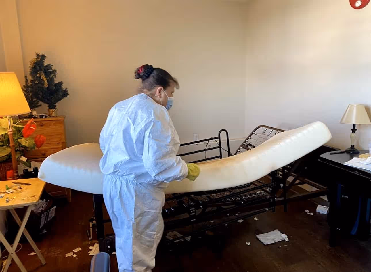 Person in protective suit and gloves cleaning a hospital bed mattress in a room with scattered papers on the floor.
