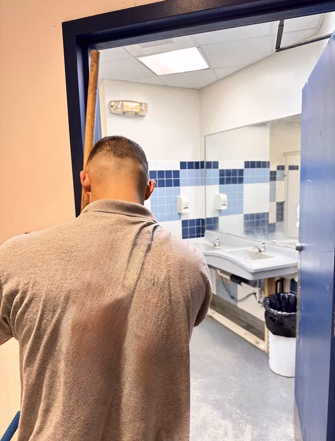 Man with short hair seen from behind holding a mop in a bathroom with blue tiled walls and a large mirror.