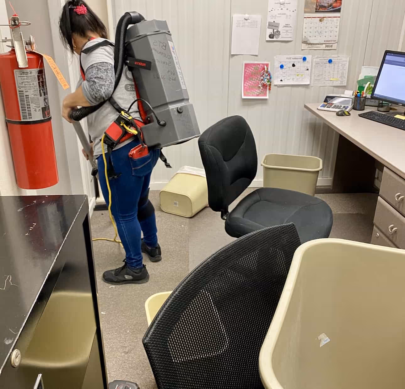 Person vacuuming the floor in an office with a backpack vacuum cleaner, surrounded by desks, chairs, and trash bins.