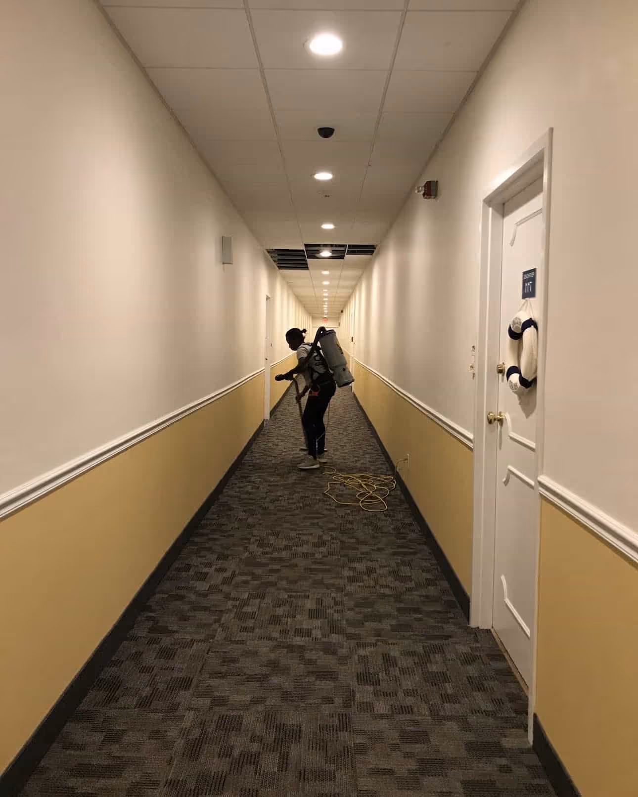 Person wearing a backpack vacuum cleaner cleaning carpet in a long, narrow hallway with beige and white walls and closed doors.