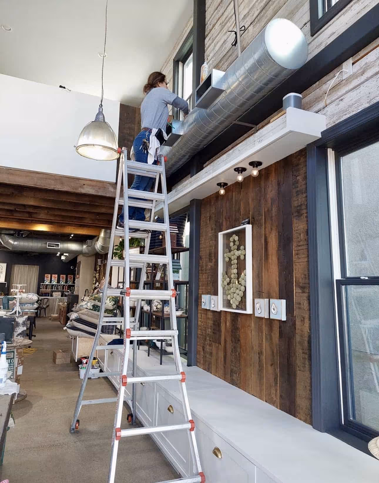 Person standing on a tall ladder working on a ventilation duct inside a spacious store with wooden and industrial decor.