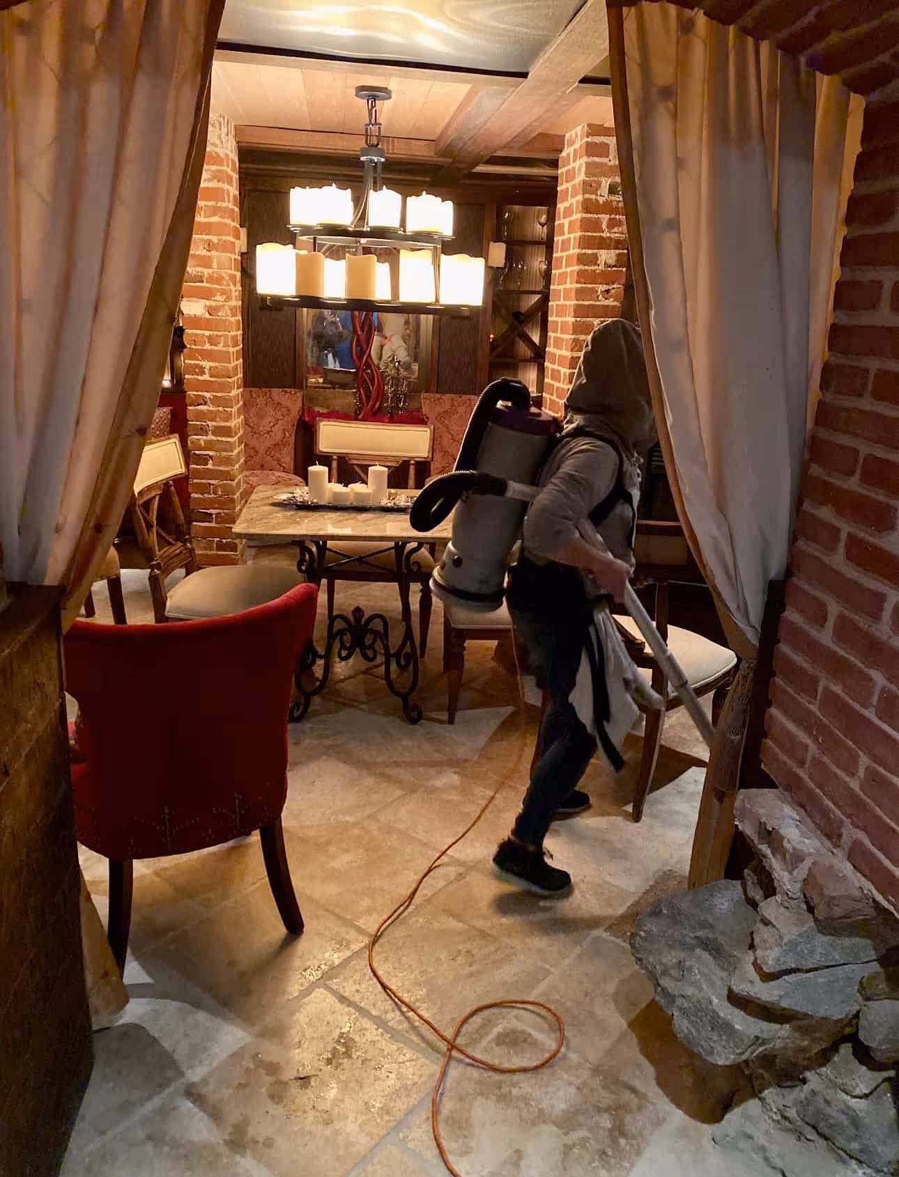 Person wearing a hood and carrying a backpack vacuum cleaner in a cozy dining room with brick walls.