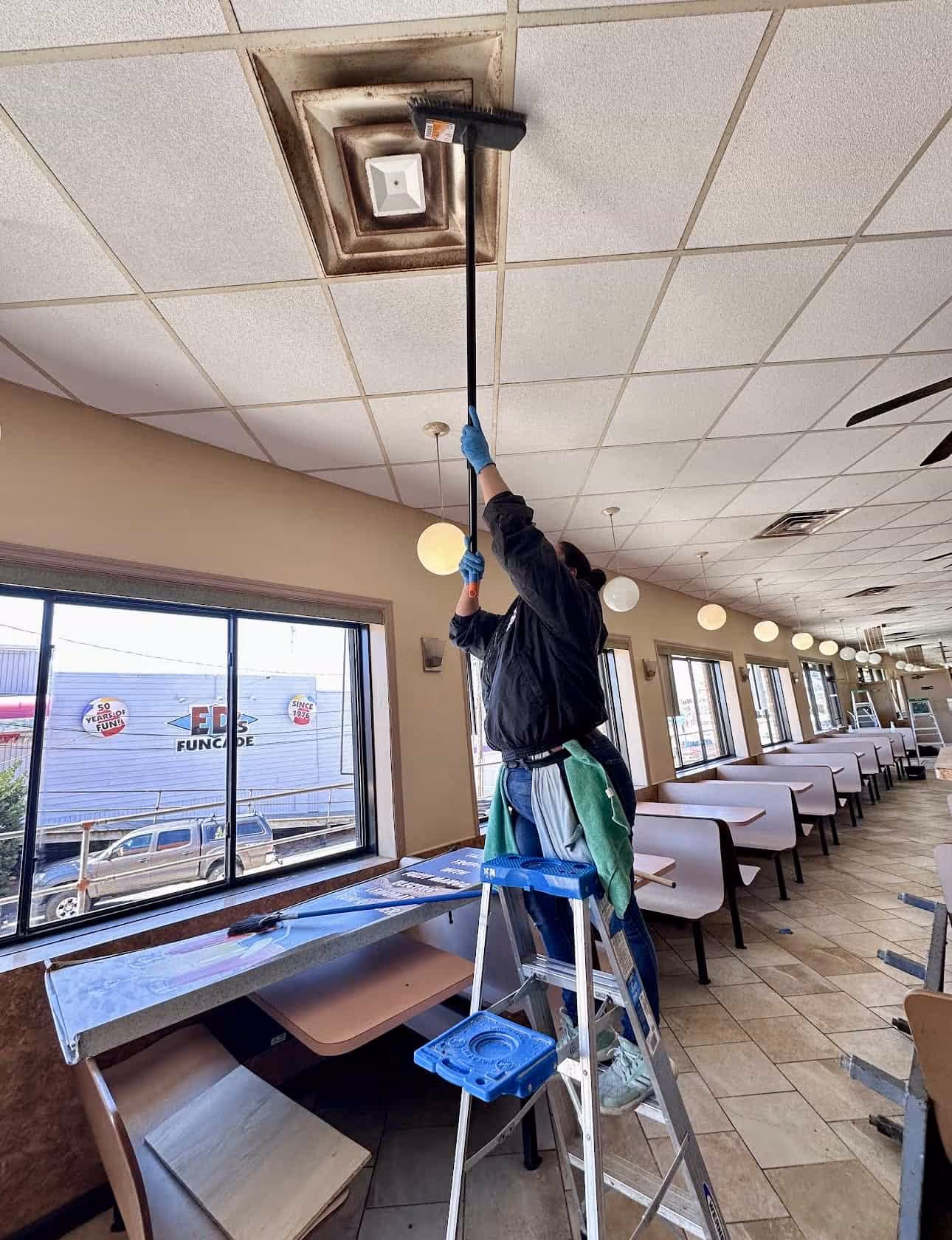 Person wearing gloves standing on a ladder cleaning a dirty ceiling vent with a long broom inside a restaurant with booths and large windows.