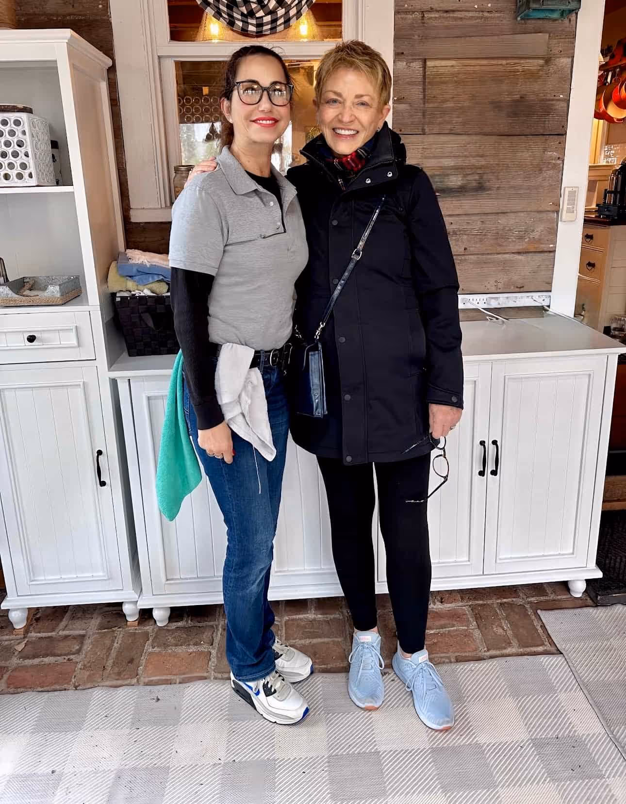 Two women standing and smiling indoors; the woman on the left wears glasses, a gray polo, jeans, and sneakers, and the woman on the right wears a black coat, black pants, blue sneakers, and holds glasses in her hand.