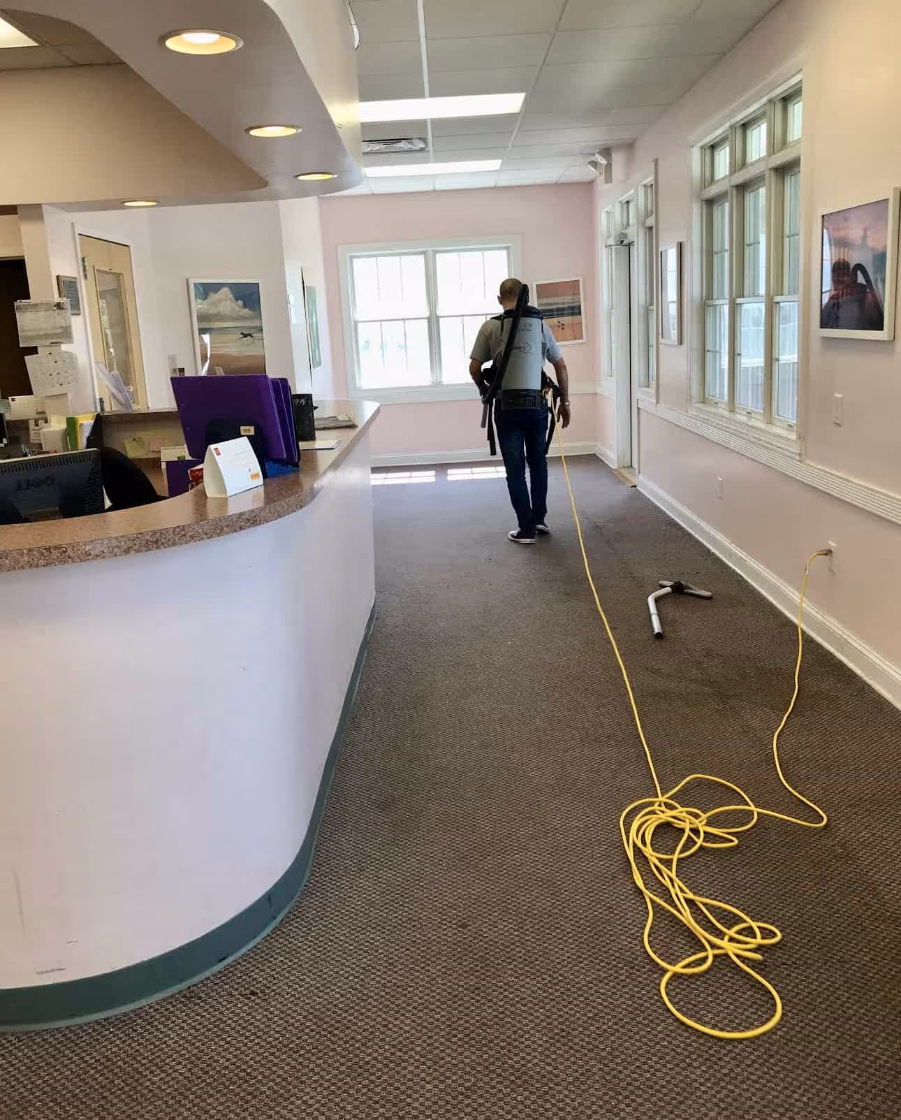 Person using a backpack vacuum cleaner to clean a carpeted office hallway with light pink walls and large windows.