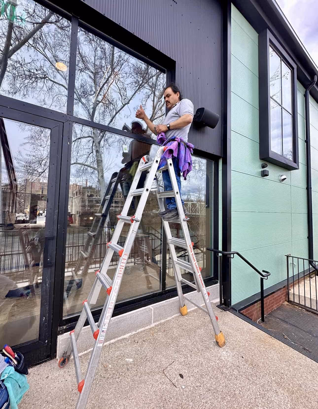 Man standing on an aluminum ladder cleaning or inspecting the large glass windows of a modern building exterior.