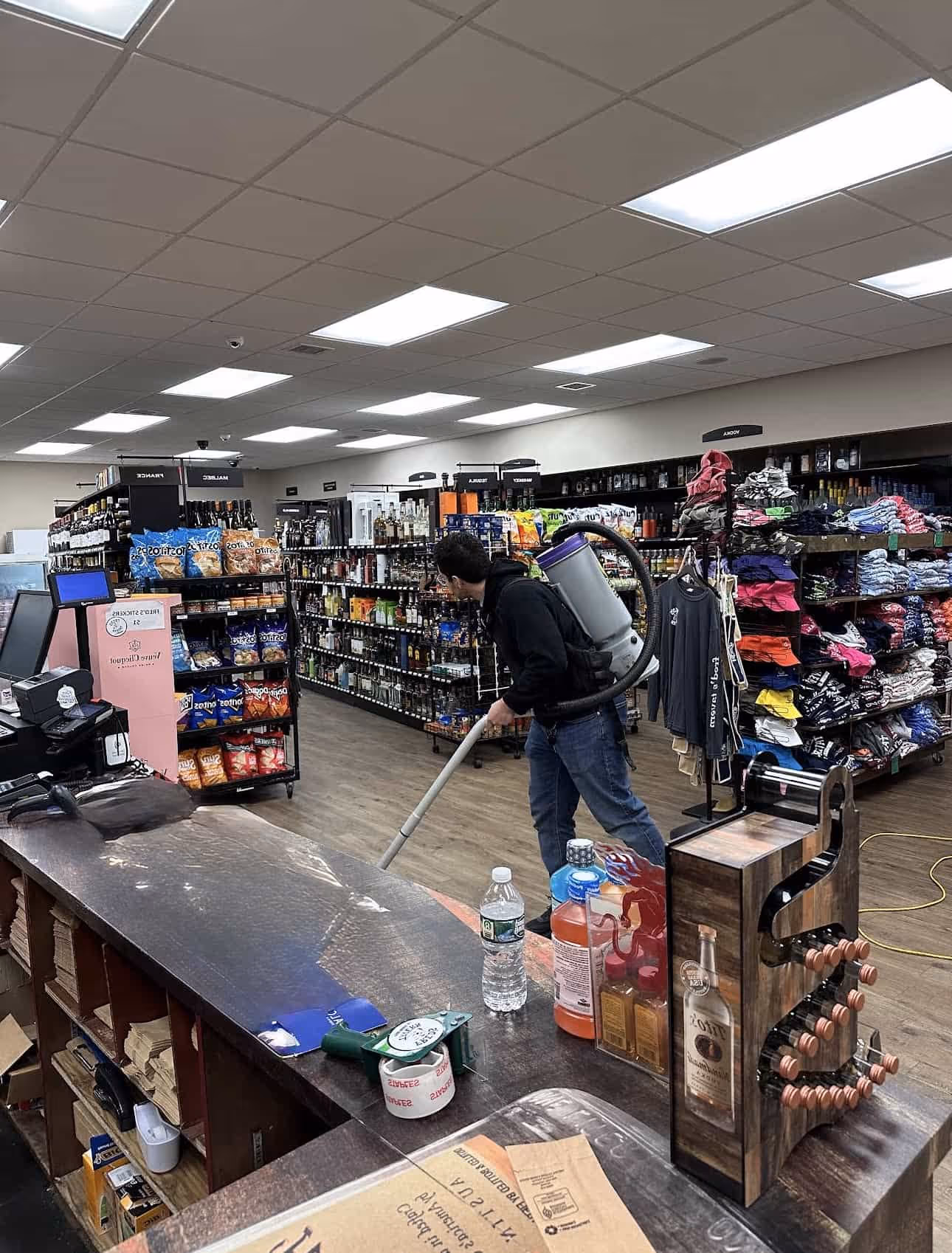 Store employee with backpack vacuum cleaner cleaning the floor near shelves stocked with chips, liquor bottles, and folded clothing.