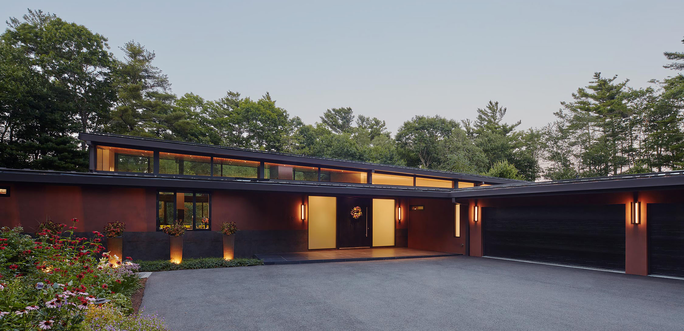 Modern single-story house with large dark garage doors, warm interior lighting, and landscaped front garden.