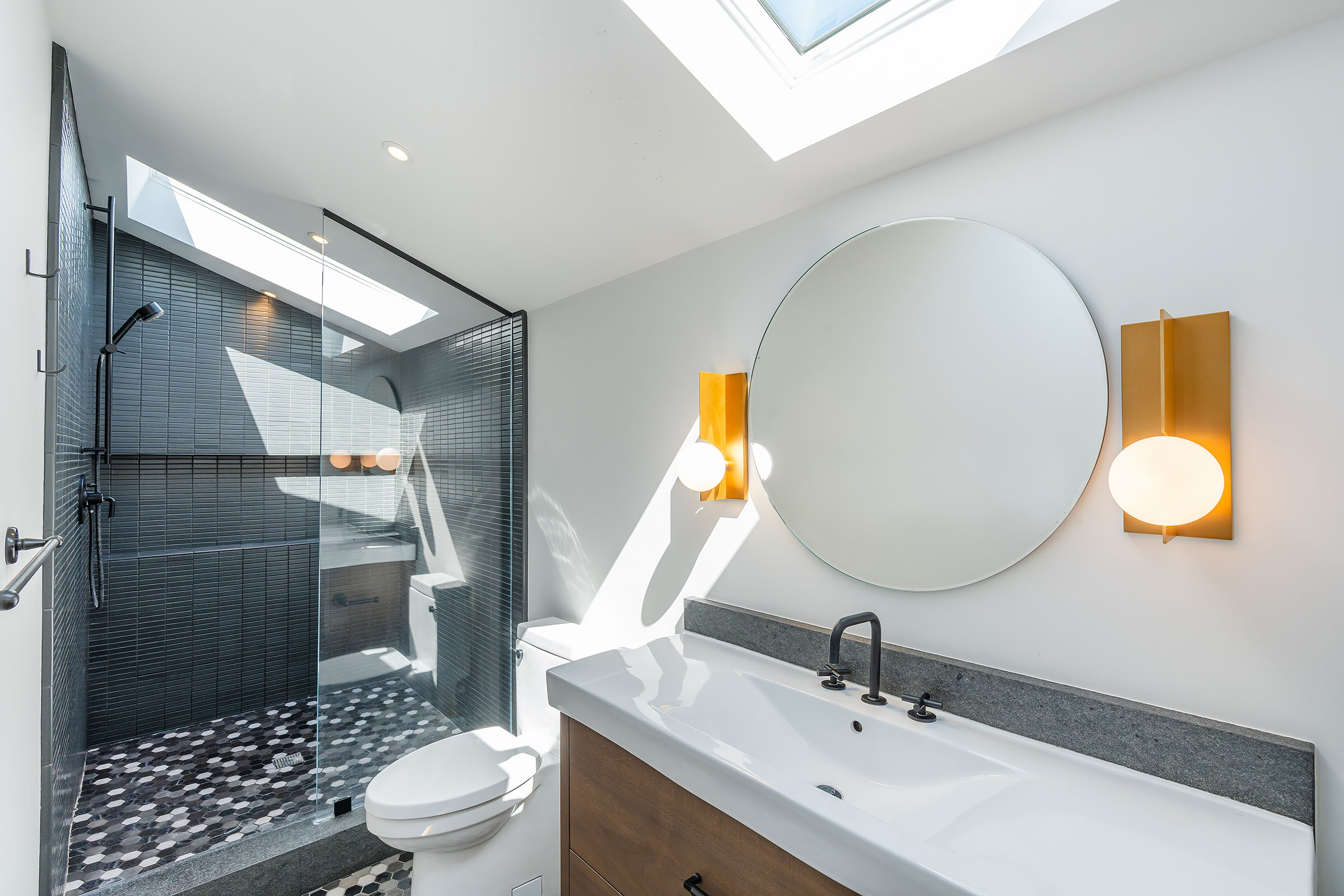 Modern bathroom with a glass-enclosed shower featuring linear and mosaic tiles, a skylit vanity with a rectangular sink, round mirror, and two wall sconces.