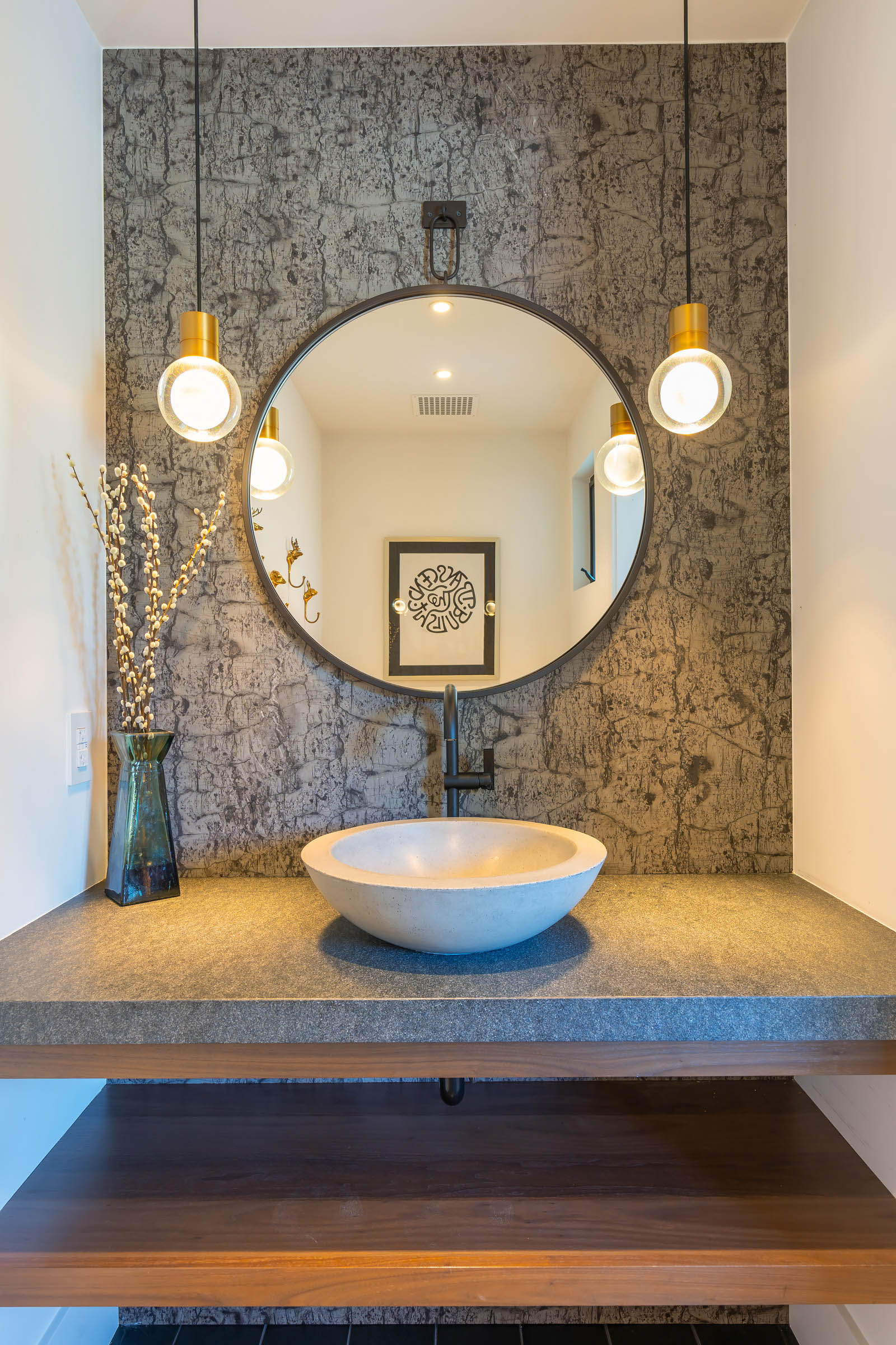 Modern powder room vanity with a round concrete vessel sink, large circular mirror, textured gray backsplash, two hanging pendant lights, and a vase with branches.