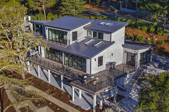 Aerial view of a modern three-story house open to terraces through expansive bifold glass doors, and surrounded by trees.