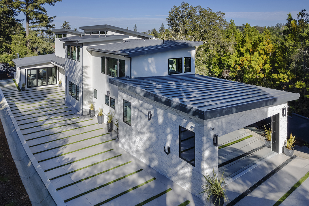 Modern two-story house with white stucco and stone exterior, large windows, metal roofing, and concrete driveway with grass inserts.