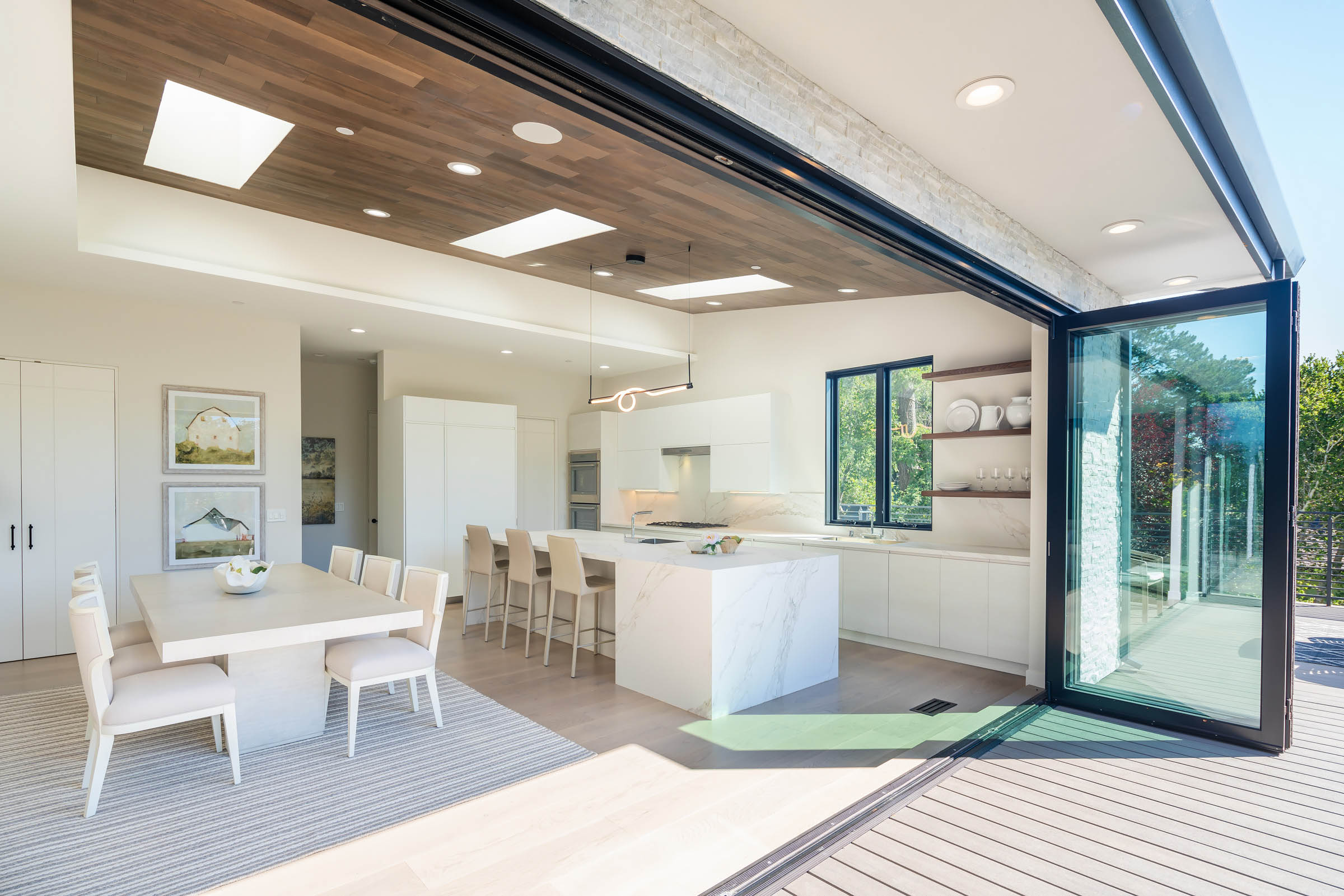 Modern open-concept kitchen and dining area with marble island, white cabinets, wooden ceiling panels, and glass doors opening to an outdoor deck.