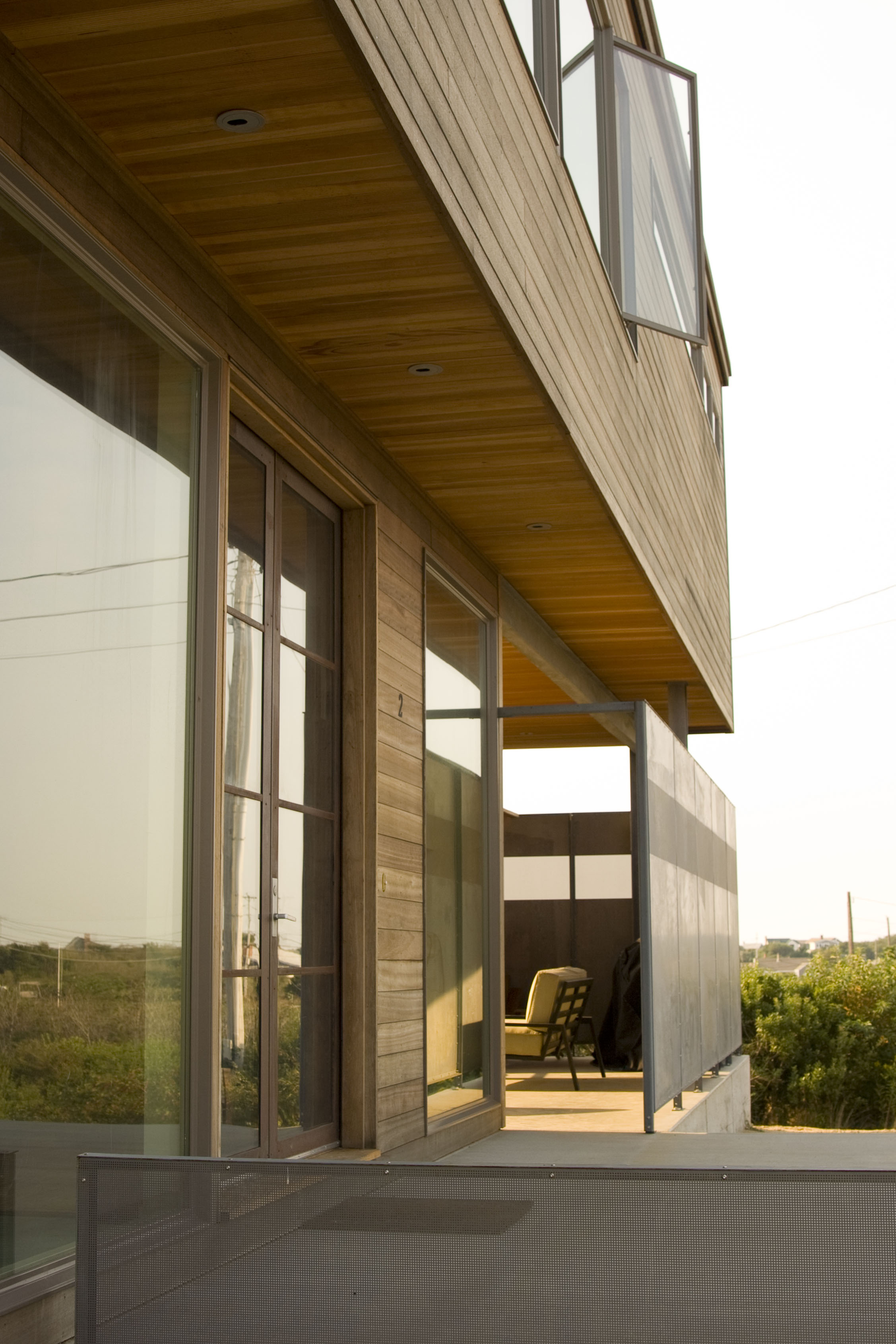 Modern house exterior with wood siding, large windows, metal panels, and covered seating area.