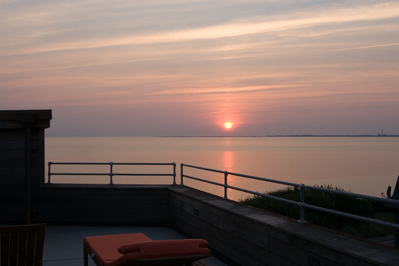 Sunset over calm ocean viewed from a roof deck with lounge chair and metal railing.