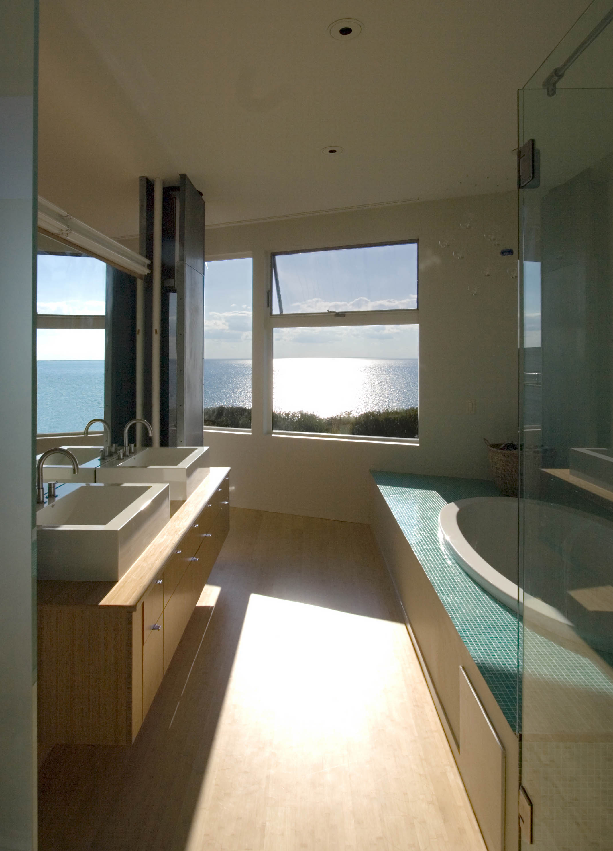 Modern bathroom with double sinks, a corner bathtub with mosaic tiles, and large windows overlooking a sunlit bay view.