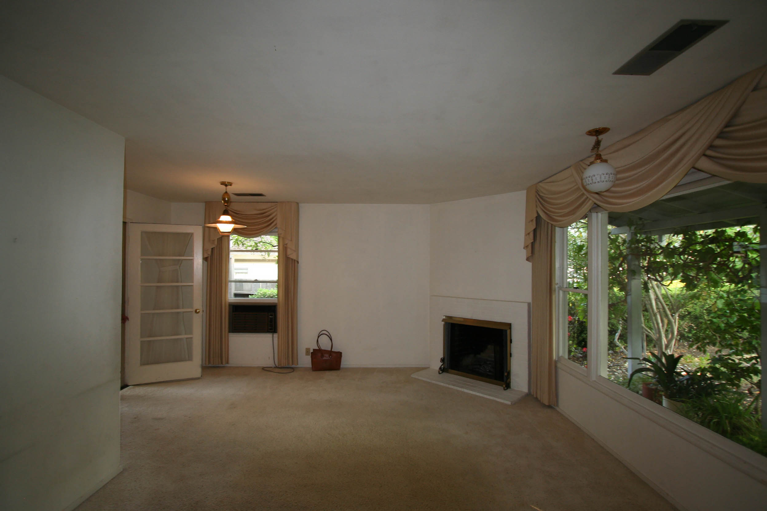 The existing living room with corner fireplace, large window with beige drapes, and a glass-paneled door.