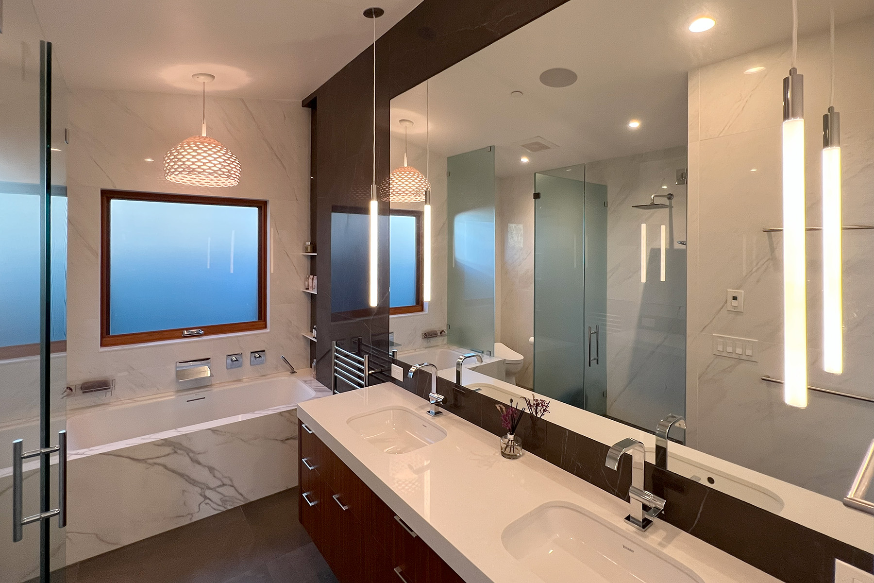 Modern bathroom with double sinks, white countertop, large mirror, floating vanity, frosted window, and bathtub with marble surround.
