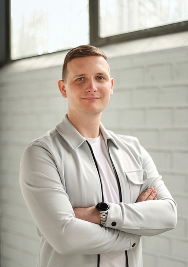 Man met korte lichtbruine haren in een lichtgrijze jas en wit T-shirt, met armen gekruist en een zwarte horloge, staand voor een raam en een lichte bakstenen muur.