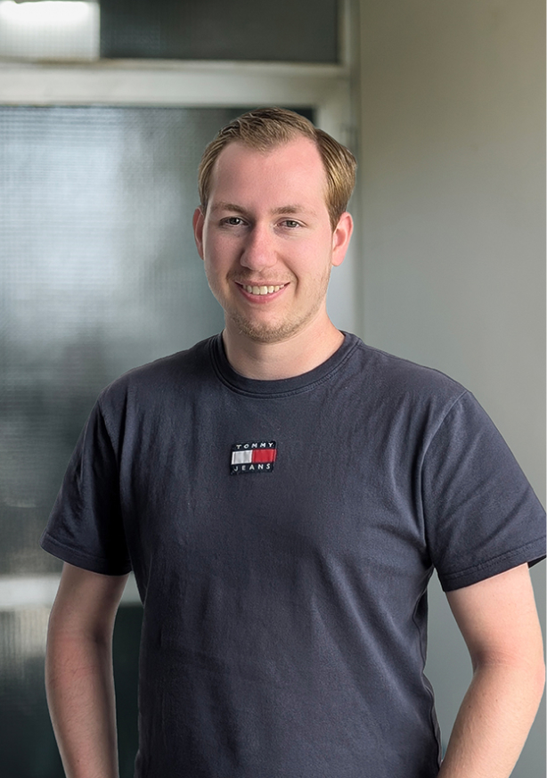 Jonge man met blond haar en een navy blauw Tommy Jeans T-shirt lacht terwijl hij voor een raam staat.