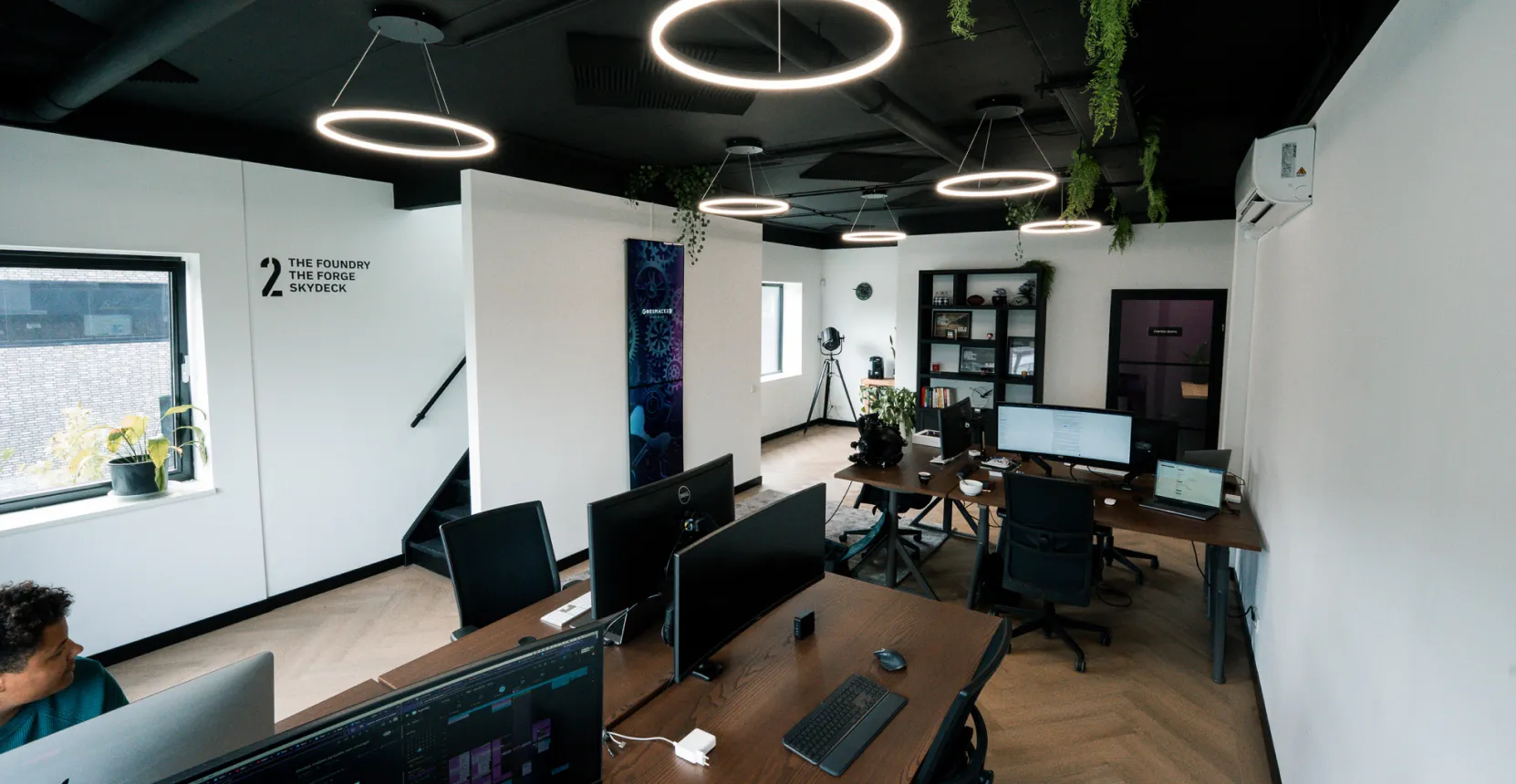 Moderne kantoorruimte met computers op houten bureaus, zwarte stoelen, ronde hanglampen en planten aan het plafond.