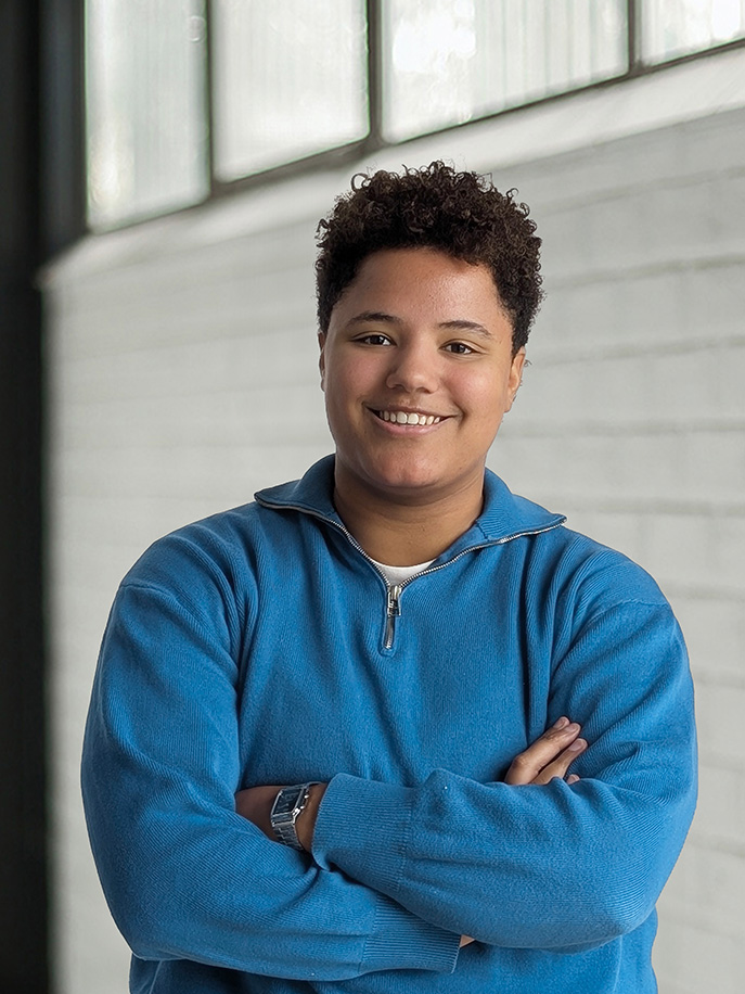 Jonge persoon met kort krullend haar draagt een blauw shirt met rits en lacht met gekruiste armen.