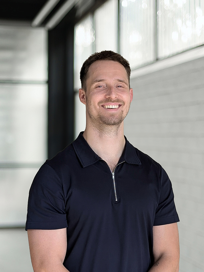 Gladde man met kort bruin haar en baard, glimlachend in een zwart shirt met korte mouwen en een ritssluiting, staand in een lichte kamer met glazen ramen.