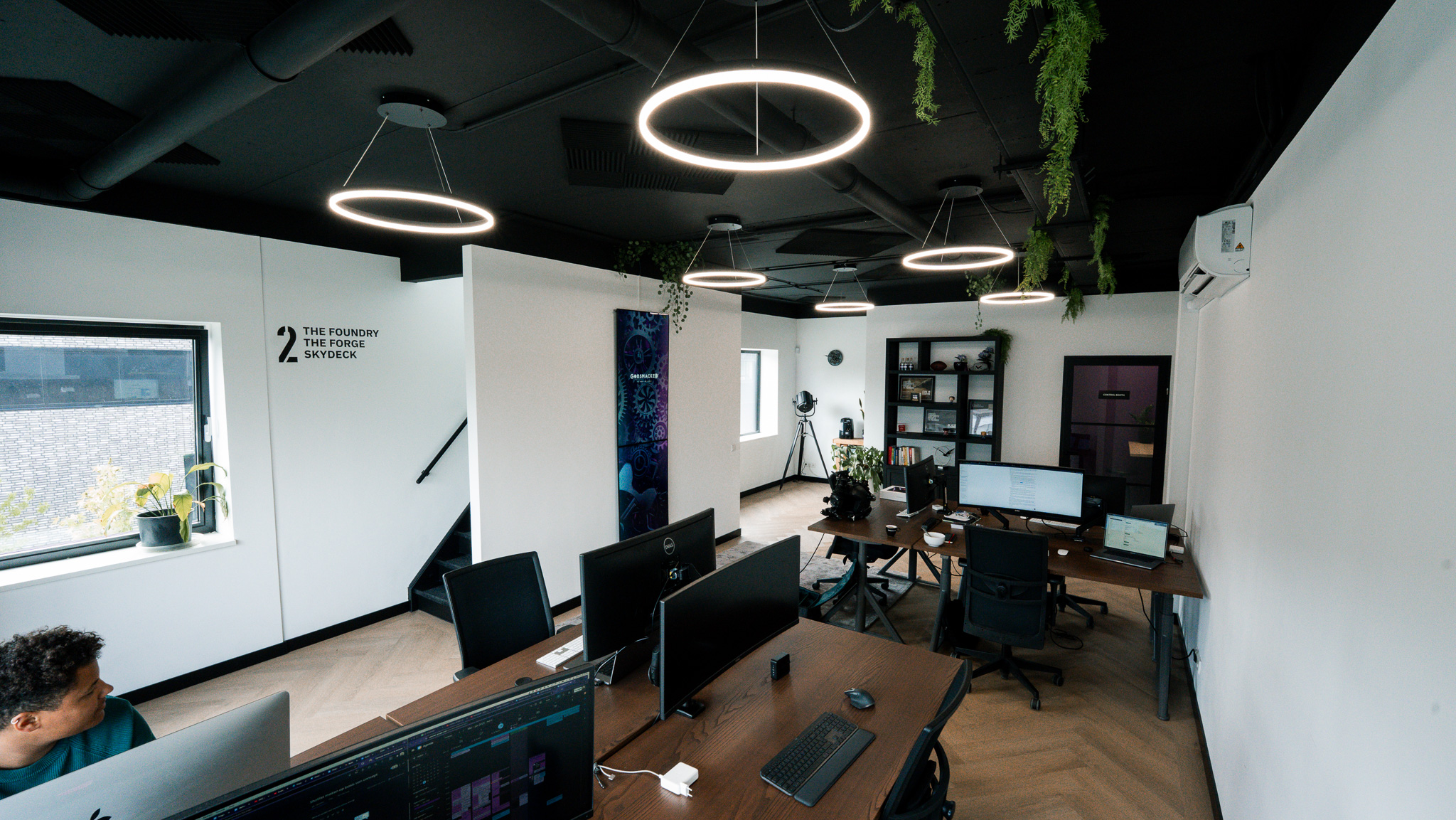 Modern kantoorruimte met houten bureaus, computerschermen, zwarte stoelen, hangende ronde ledlampen en planten, onder een zwart plafond.