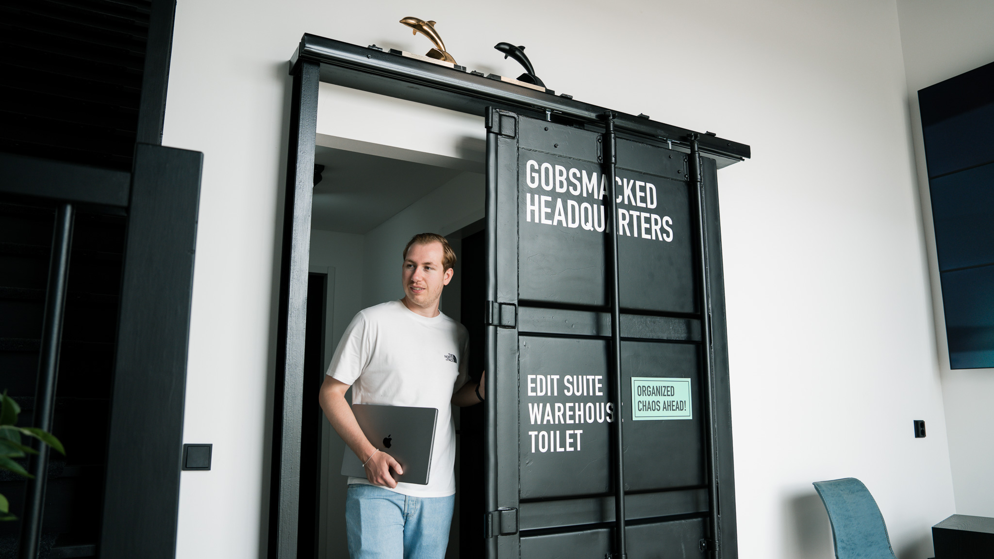 Man met een laptop die een schuifdeur opent met de tekst 'Gobsmacked Headquarters', 'Edit Suite', 'Warehouse', 'Toilet' en een bord met 'Organized Chaos Ahead!'