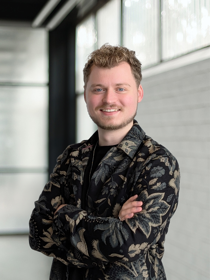 Jonge man met lichte krullende haren en baard, glimlachend met armen gekruist, draagt een zwart overhemd met een bloemmotief, staande in een lichte, moderne binnenruimte.