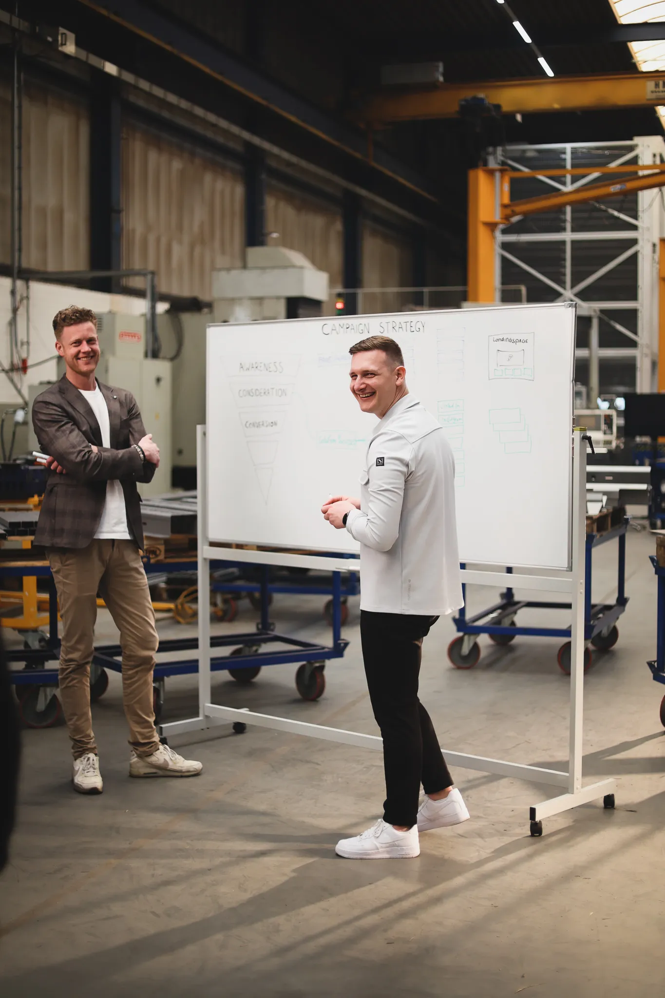 Twee mannen staan lachend in een industriële ruimte voor een whiteboard met daarop een campagne strategie en bijbehorende diagrammen.