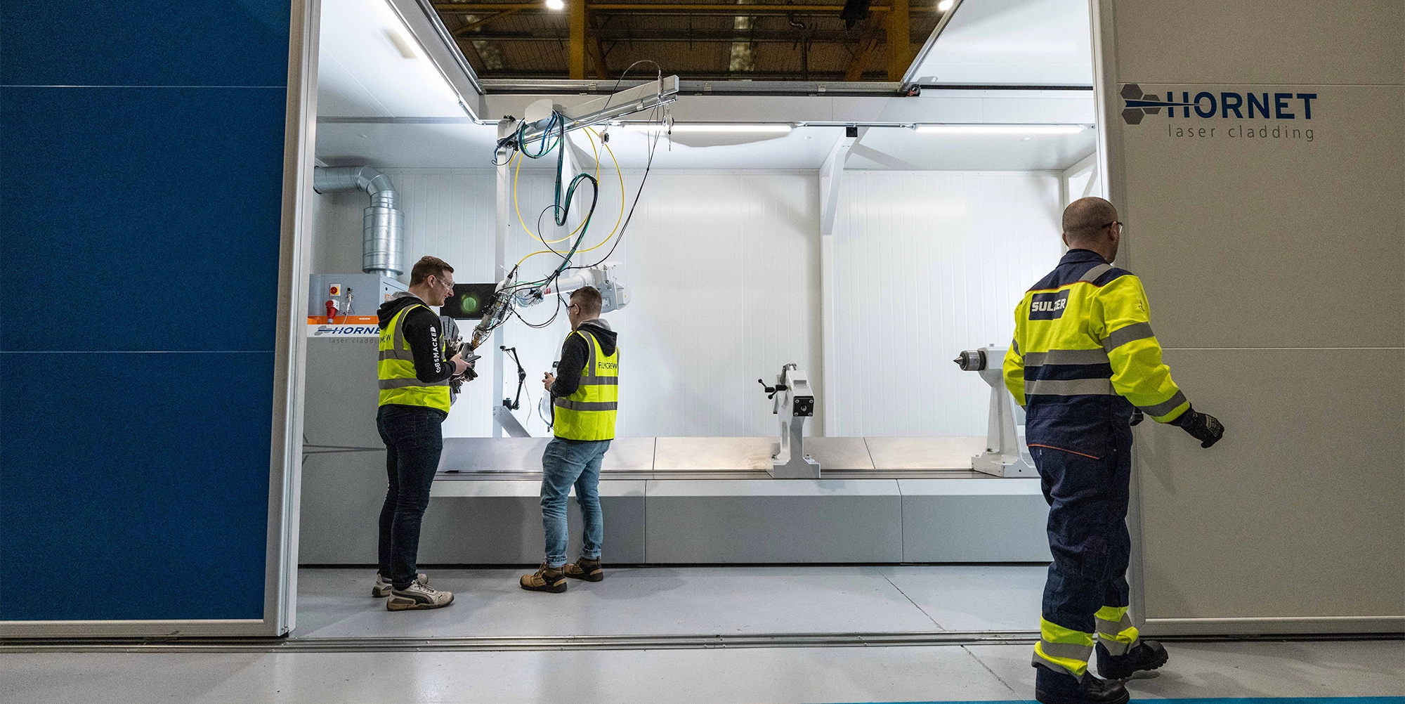 Drie werknemers in veiligheidskleding bestuderen een laserlageringsmachine in een industriële werkruimte.