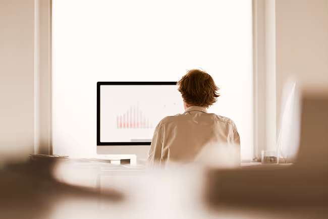 Person with short hair working on a desktop computer displaying a bar graph in a bright office.