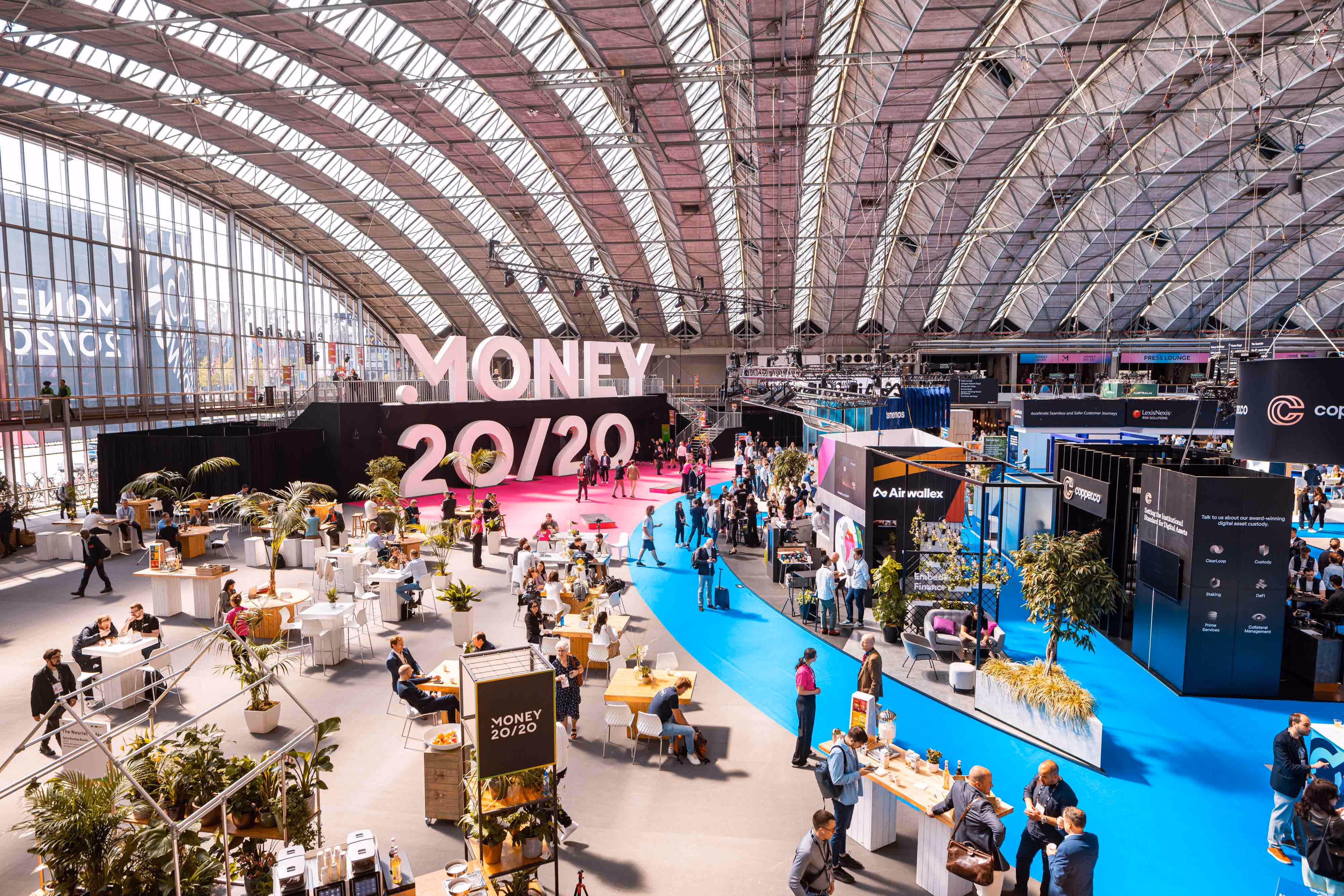 Large indoor event space with curved glass ceiling hosting the MONEY 20/20 conference, featuring people walking and sitting at tables, colorful blue and pink carpeted areas, and various booths and displays.