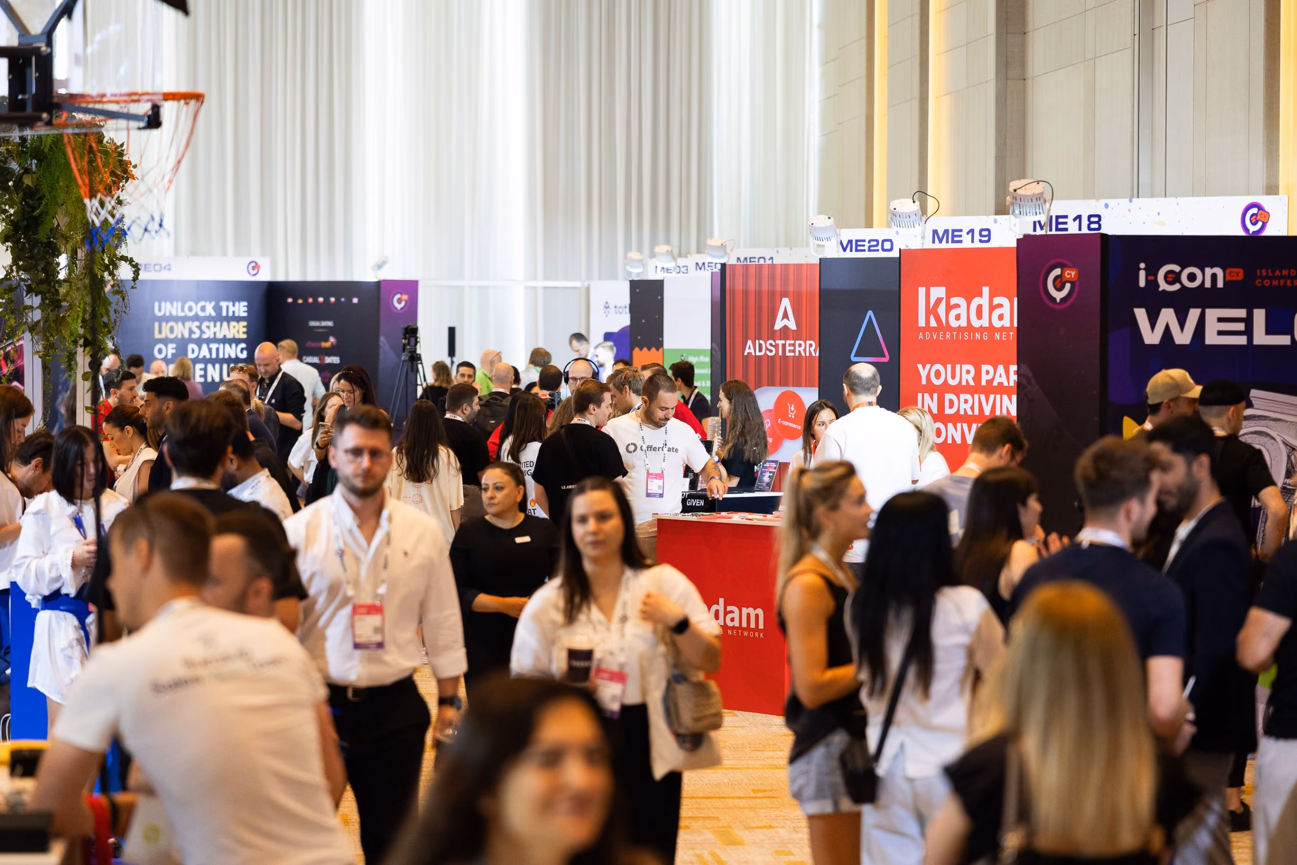 Crowd of people walking and interacting at an indoor event with advertising booths and banners, including visible signs for 'Adsterra' and 'Kadam'.