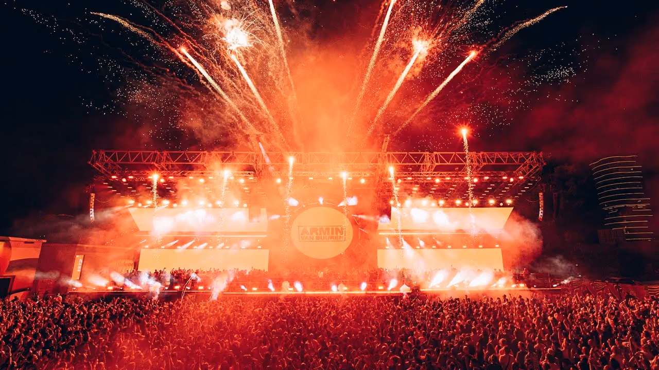 Large crowd at a nighttime outdoor concert with a brightly lit stage and fireworks exploding overhead.
