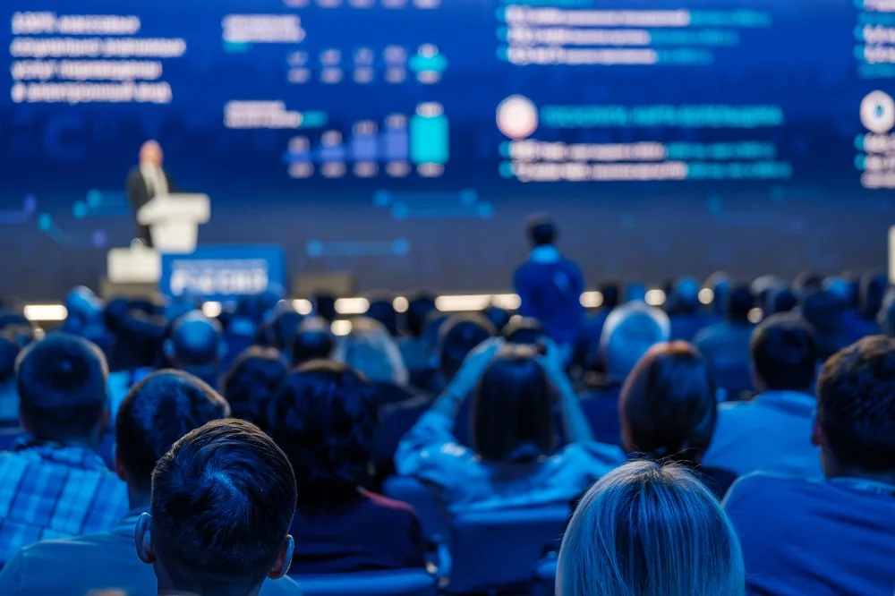 Audience seated and listening to a speaker giving a presentation on stage with a large screen displaying charts and text in the background at a conference.