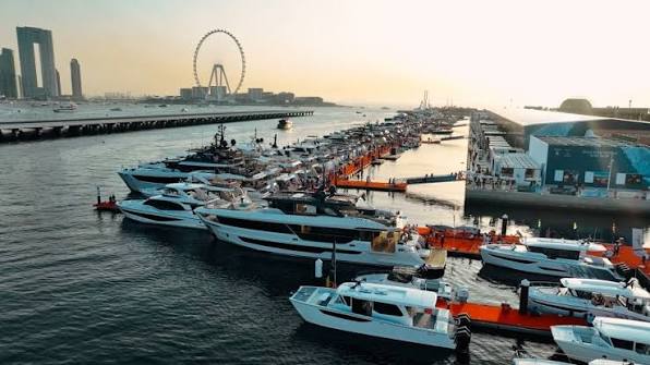 A marina at sunset with multiple yachts and boats docked along piers, a large Ferris wheel and tall buildings in the background under a clear sky.