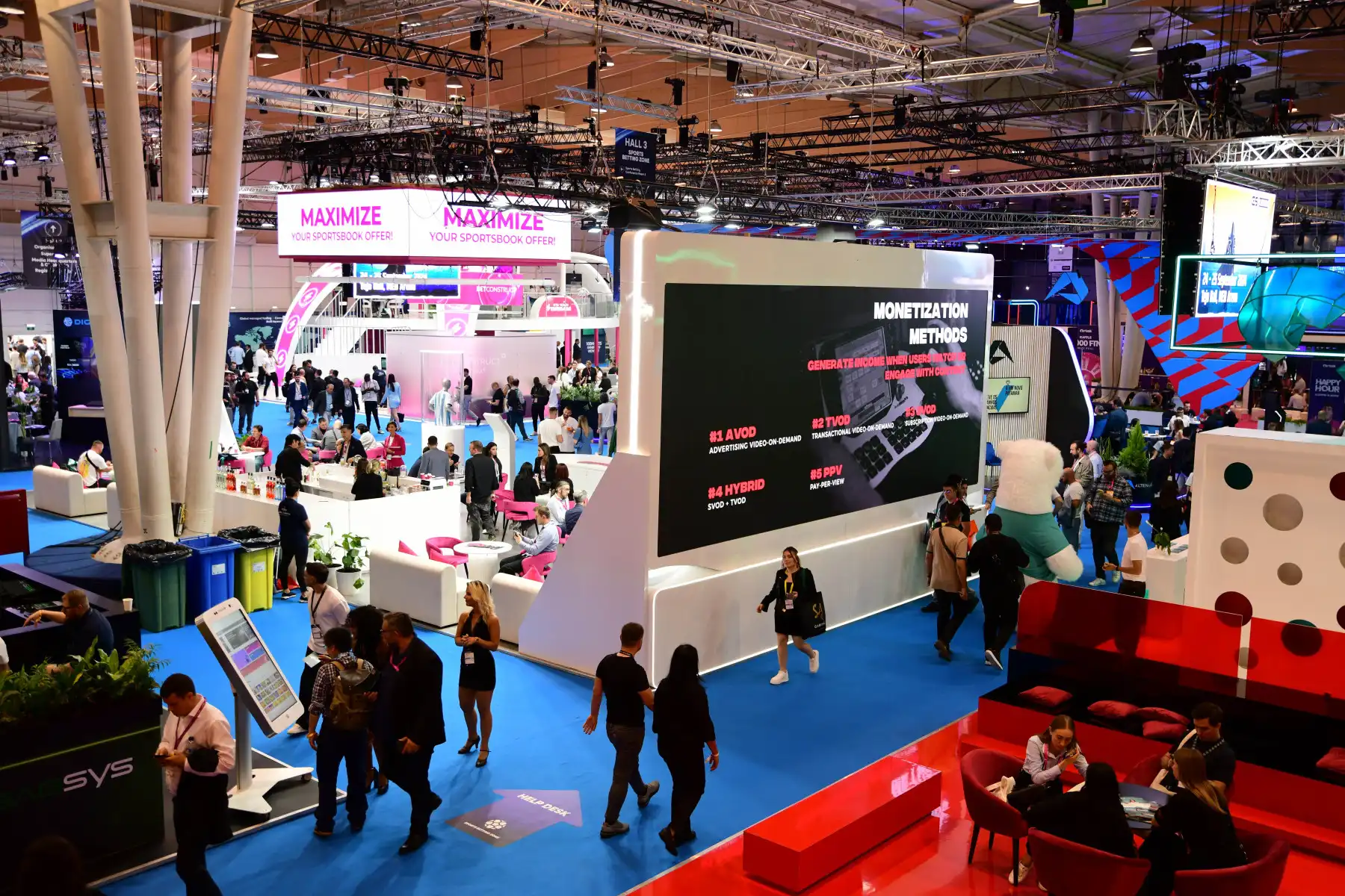 Wide view of a busy trade show or conference with attendees walking and sitting in booths, a large digital screen displaying monetization methods like AVOD, TVOD, SVOD, and PPV, colorful seating areas, and promotional signs including a pink sign that says MAXIMIZE YOUR SPORTSBOOK OFFER.
