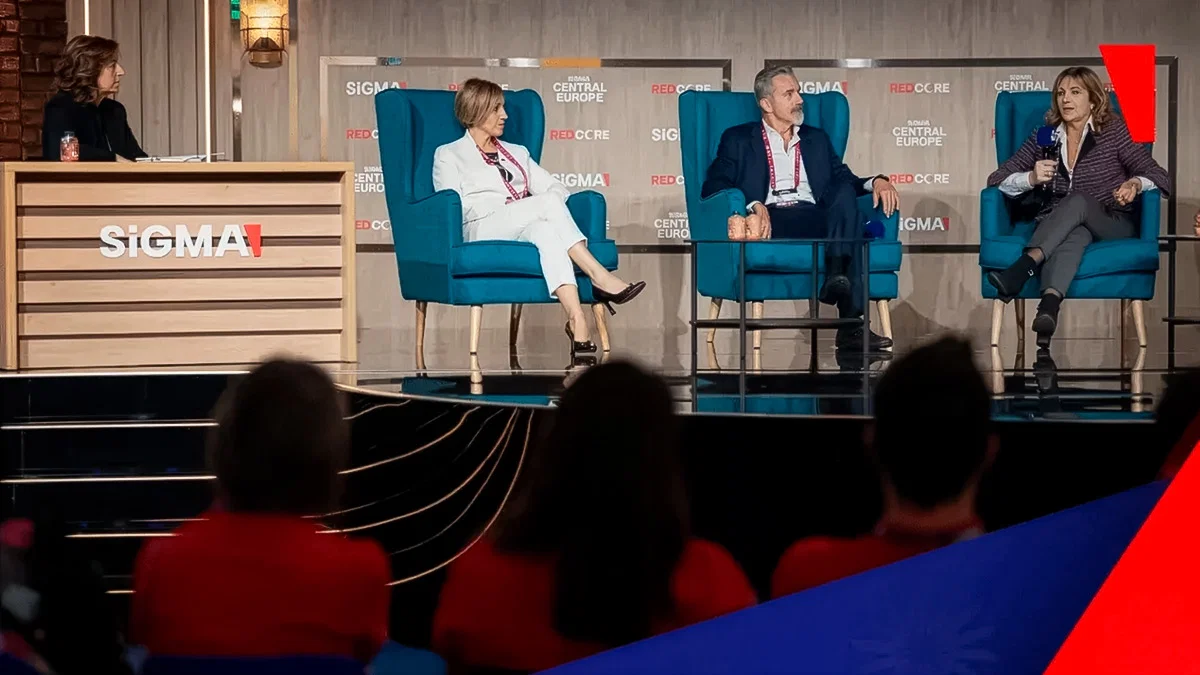 Panel discussion at a conference with four speakers: one woman at a wooden podium labeled 'SiGMA' and three seated panelists conversing in blue armchairs on stage, facing an audience.