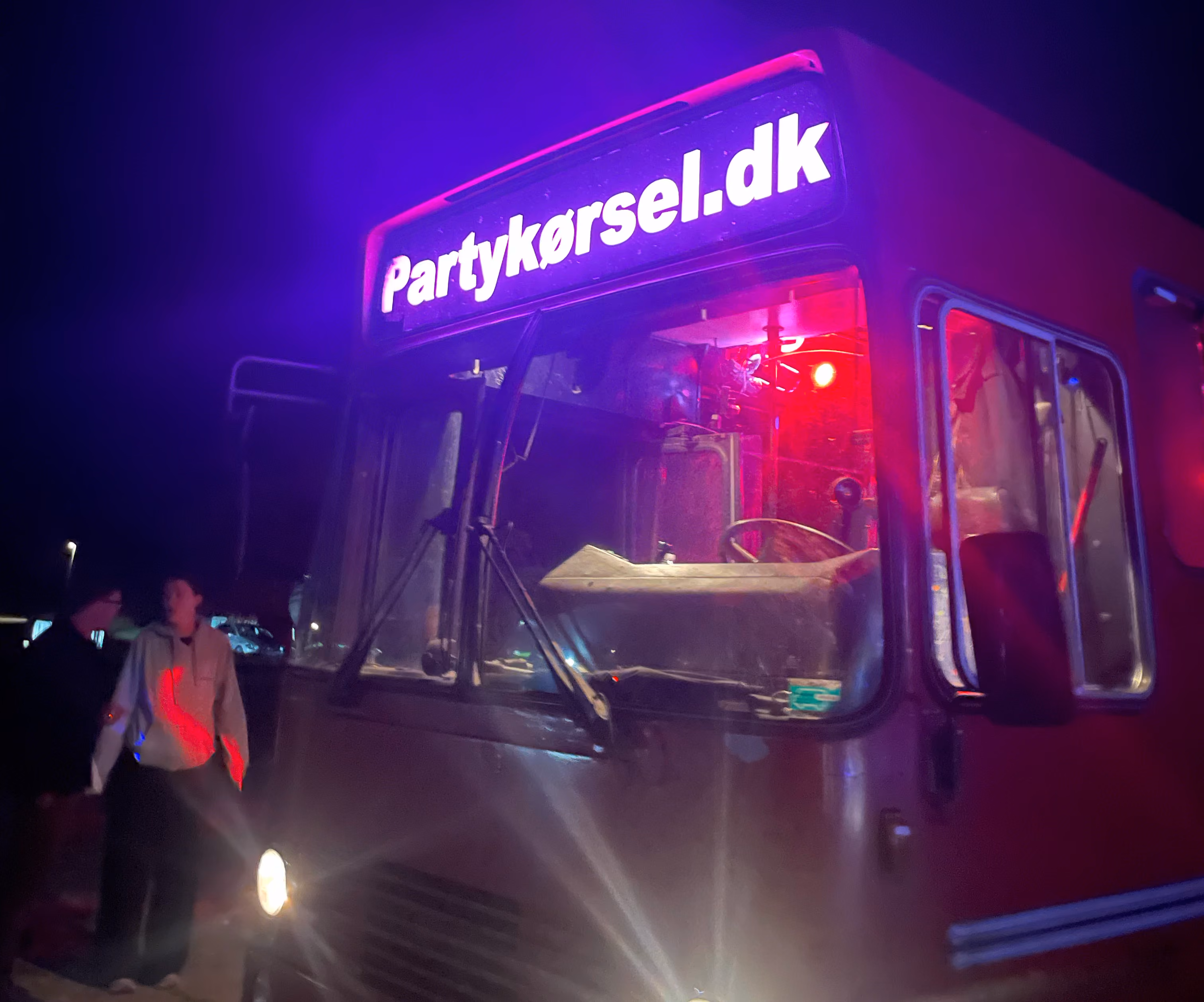 Rød bus med oplyst skilt 'Partykørsel.dk' og to personer stående ved siden af under nattelys.