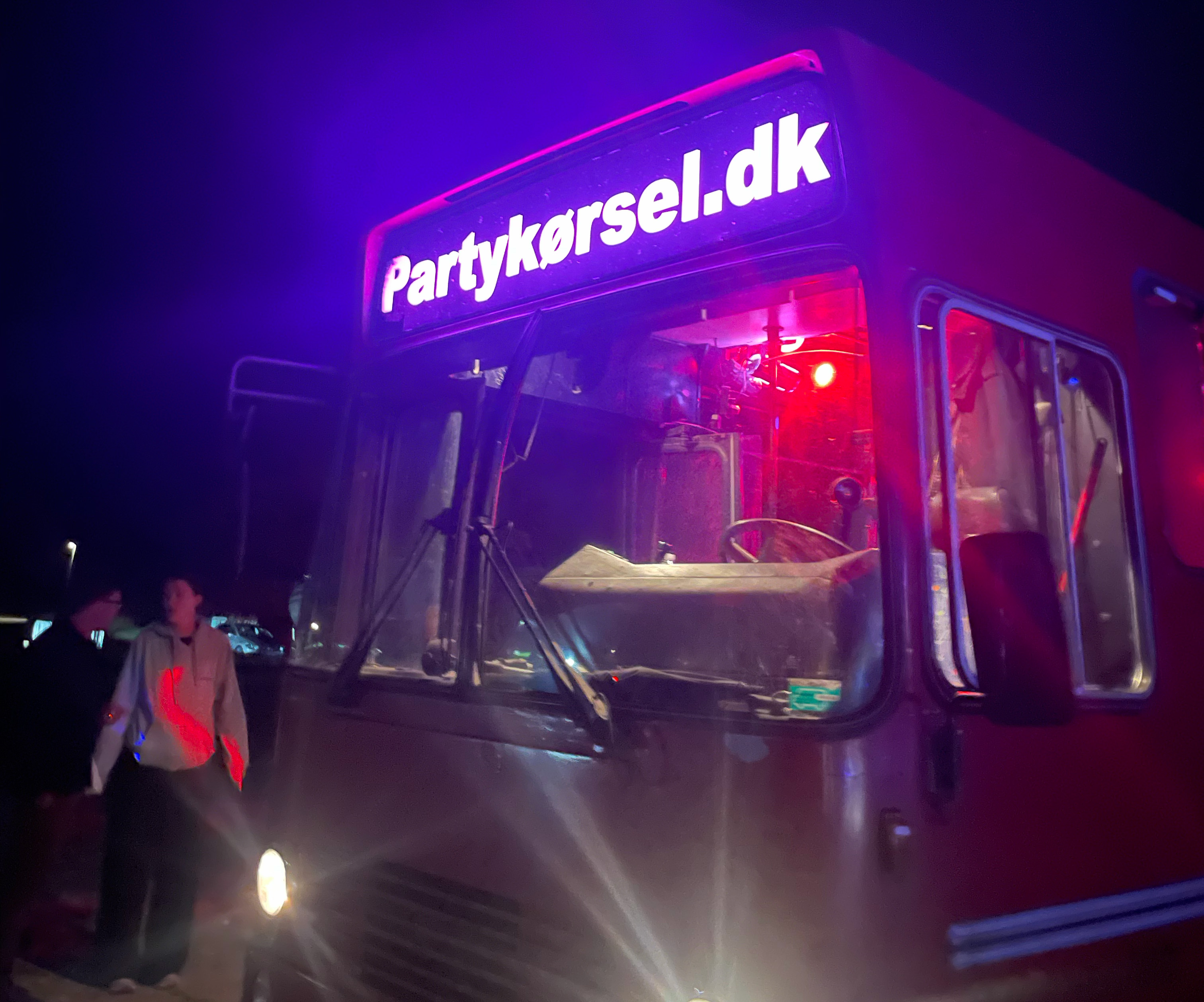 Rød bus med oplyst skilt 'Partykørsel.dk' og to personer stående ved siden af under nattelys.