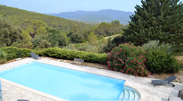 Piscine rectangulaire extérieure avec eau bleu clair, entourée de pavés en pierre, de chaises longues et de buissons verts, avec collines et montagnes en arrière-plan.