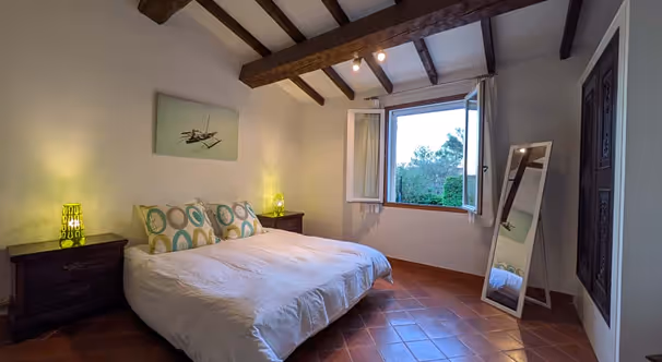 Chambre chaleureuse avec poutres en bois au plafond, lit double avec coussins à motifs, deux tables de chevet avec lampes, fenêtre ouverte et miroir sur pied.